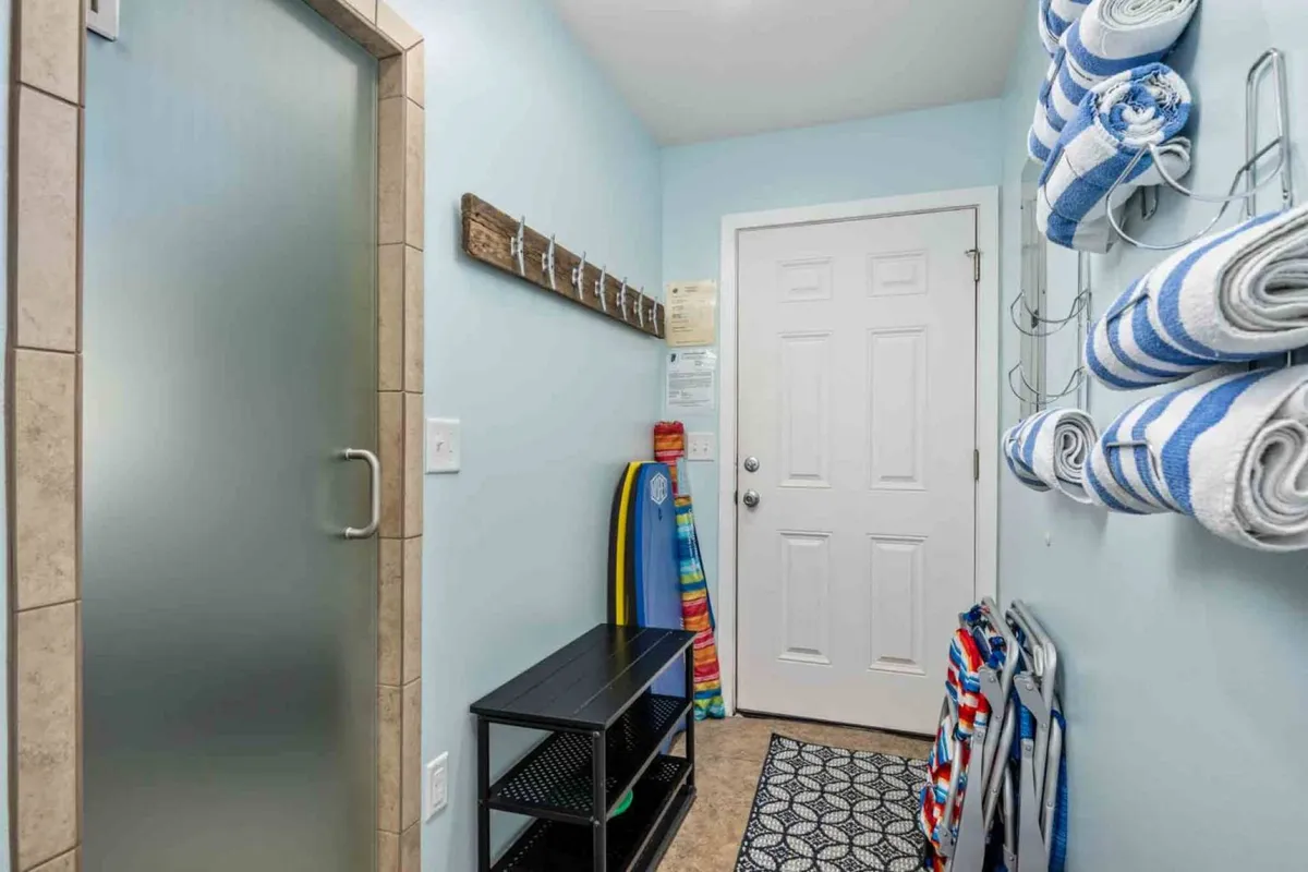 A functional and beach-inspired space with a wooden coat rack for leashes and towels, a black storage unit for pet or beach gear, and surfboards propped against the wall. Colorful towels add a playful touch, while the frosted shower door hints at easy cleanup after outdoor adventures.