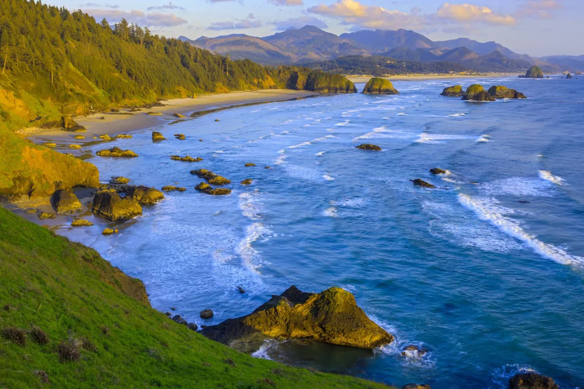 A sweeping view of the coastline, featuring a sandy beach, gentle ocean waves, striking rock formations, and distant mountains under a serene sky. The image captures the natural beauty and tranquility of the area, inviting viewers to imagine themselves exploring the outdoors.
