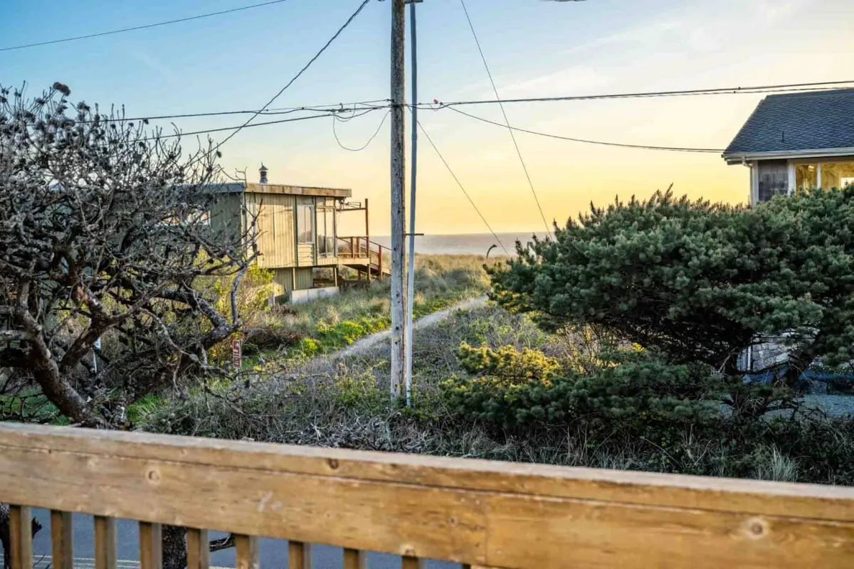 This image captures a peaceful pathway leading directly to the sandy beach, framed by surrounding trees, a distant ocean, and a glowing sunset sky. The view hints at nearby houses, suggesting a convenient and inviting connection from the home to the beach. The tranquil mood is highlighted by soft evening light and the inviting natural setting.