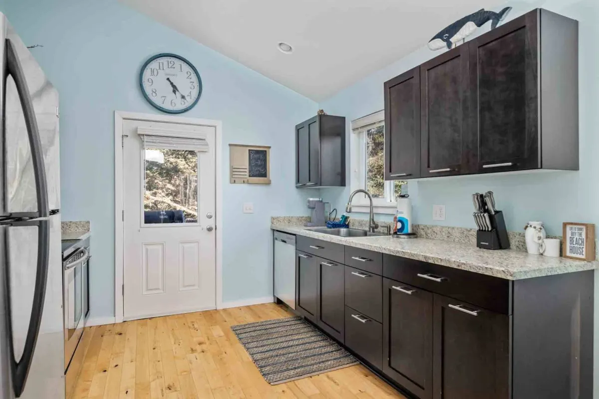 A welcoming kitchen interior featuring granite countertops, dark cabinetry, a window that frames an outdoor view, and a traditional wall clock. The space is well-lit, inviting, and perfectly suited for cooking and entertaining, with subtle touches that suggest family gatherings and holiday meals.