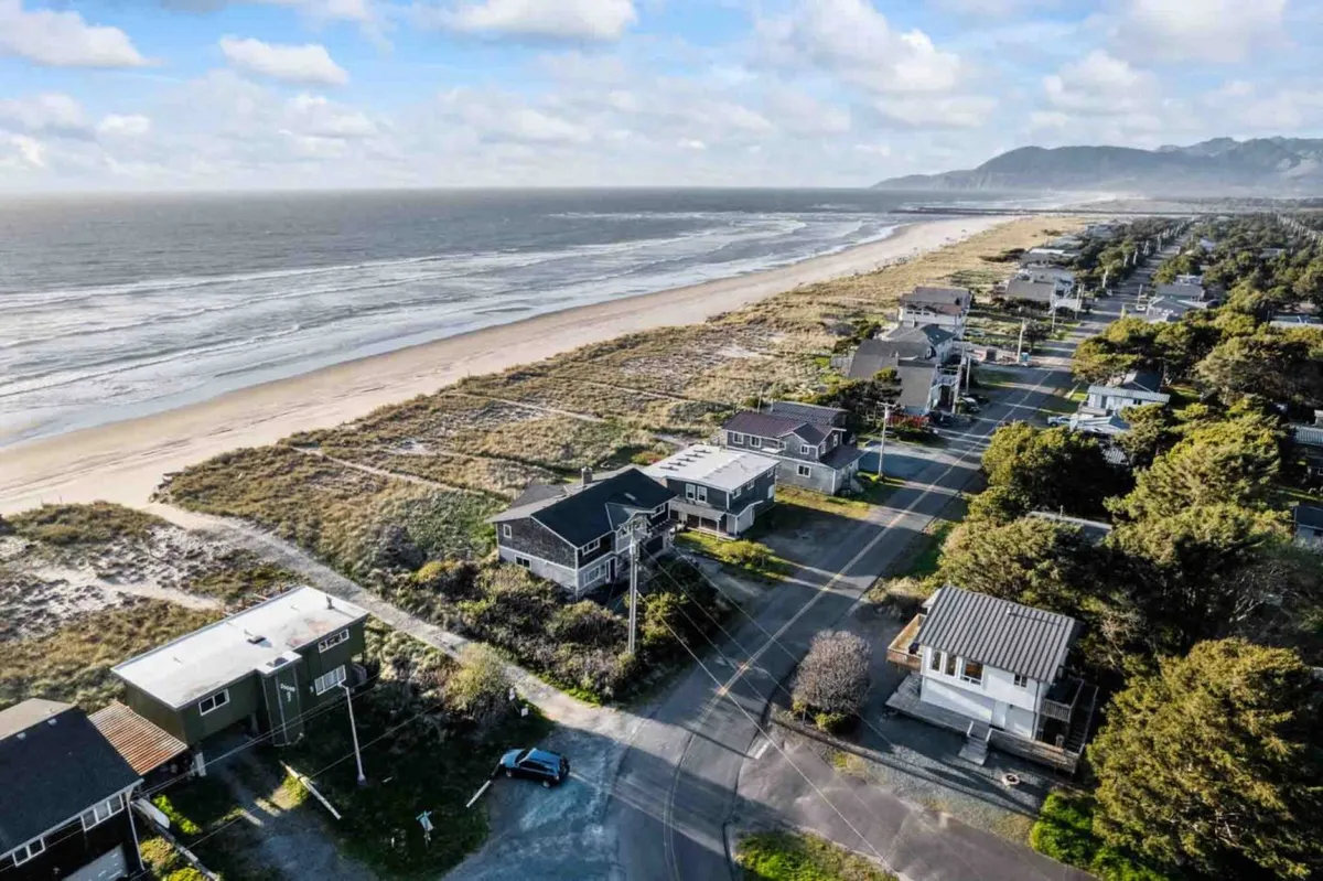 The hero image displays a breathtaking view of the wide, sandy beach stretching alongside the tranquil ocean waters, with charming coastal houses and a quiet road nestled just steps from the shore. The scene exudes a relaxed mood, perfect for sunset strolls, surfing, fishing, and soaking in the serene coastal atmosphere.