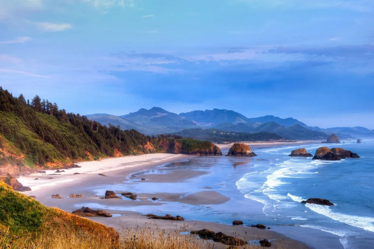 A sweeping view of Nedonna Beach’s wide sandy expanse, with gentle waves rolling ashore, dramatic rock formations, and a distant mountain range beneath a vast sky. The tranquil atmosphere invites beachgoers to walk, explore, and relax in nature’s embrace.