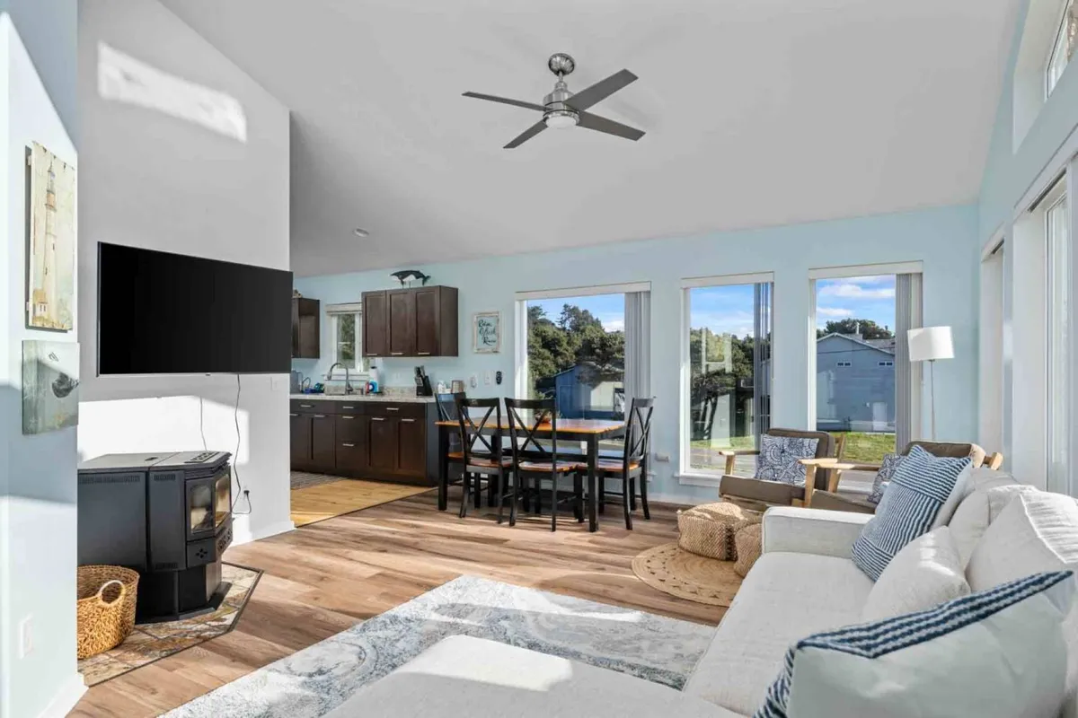 This interior view shows a cozy living space with a sofa featuring decorative cushions, a dining table nearby, and expansive windows letting in sunlight. The wooden flooring and overall layout create a seamless transition between lounging and dining areas. The windows reveal glimpses of the outside, hinting at the impressive views beyond.