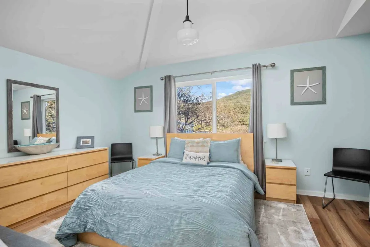 A serene bedroom featuring a bed with light blue bedding, a large window offering a scenic view, decorative starfish art above the bed, and a modern dresser. Natural light fills the room, giving it a peaceful, airy atmosphere.